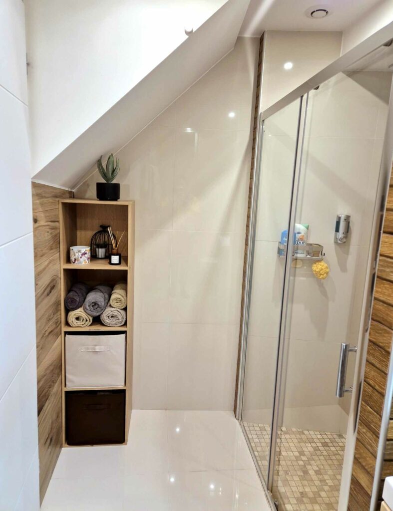 Espace douche avec paroi vitrée, étagère en bois contenant des serviettes roulées et des paniers de rangement