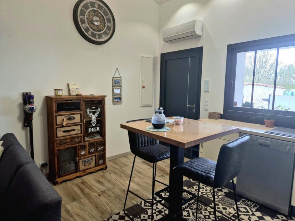 Vue d'un intérieur aménagé comprenant une table haute en bois avec deux chaises de bar noires sur un tapis sol carreaux de ciment. À gauche, un buffet en bois de style apothicaire avec des tiroirs dépareillés et une horloge murale industrielle. À droite, un plan de travail de cuisine avec lave-vaisselle intégré sous une fenêtre, le tout dans une pièce aux murs blancs et sol en imitation bois.