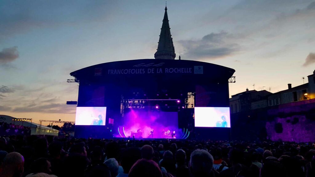 Scène de concert illuminée en violet sous un ciel de crépuscule aux Francofolies de La Rochelle avec une foule compacte