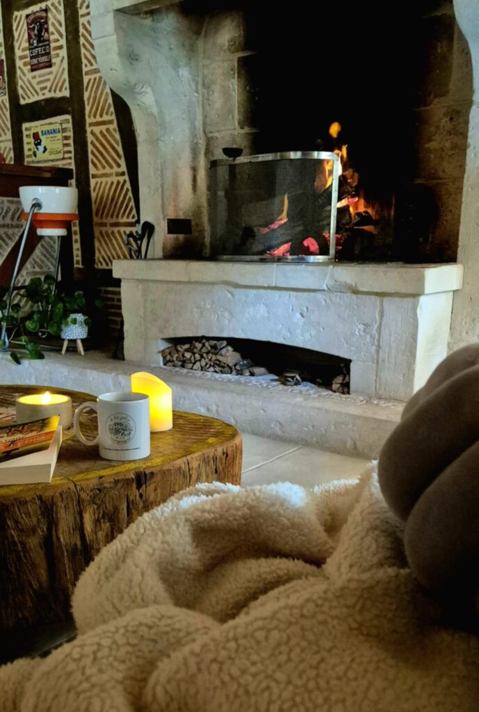 Un salon chaleureux avec un feu de cheminée crépitant, une tasse posée sur une table en bois brut, des bougies allumées et un plaid douillet au premier plan