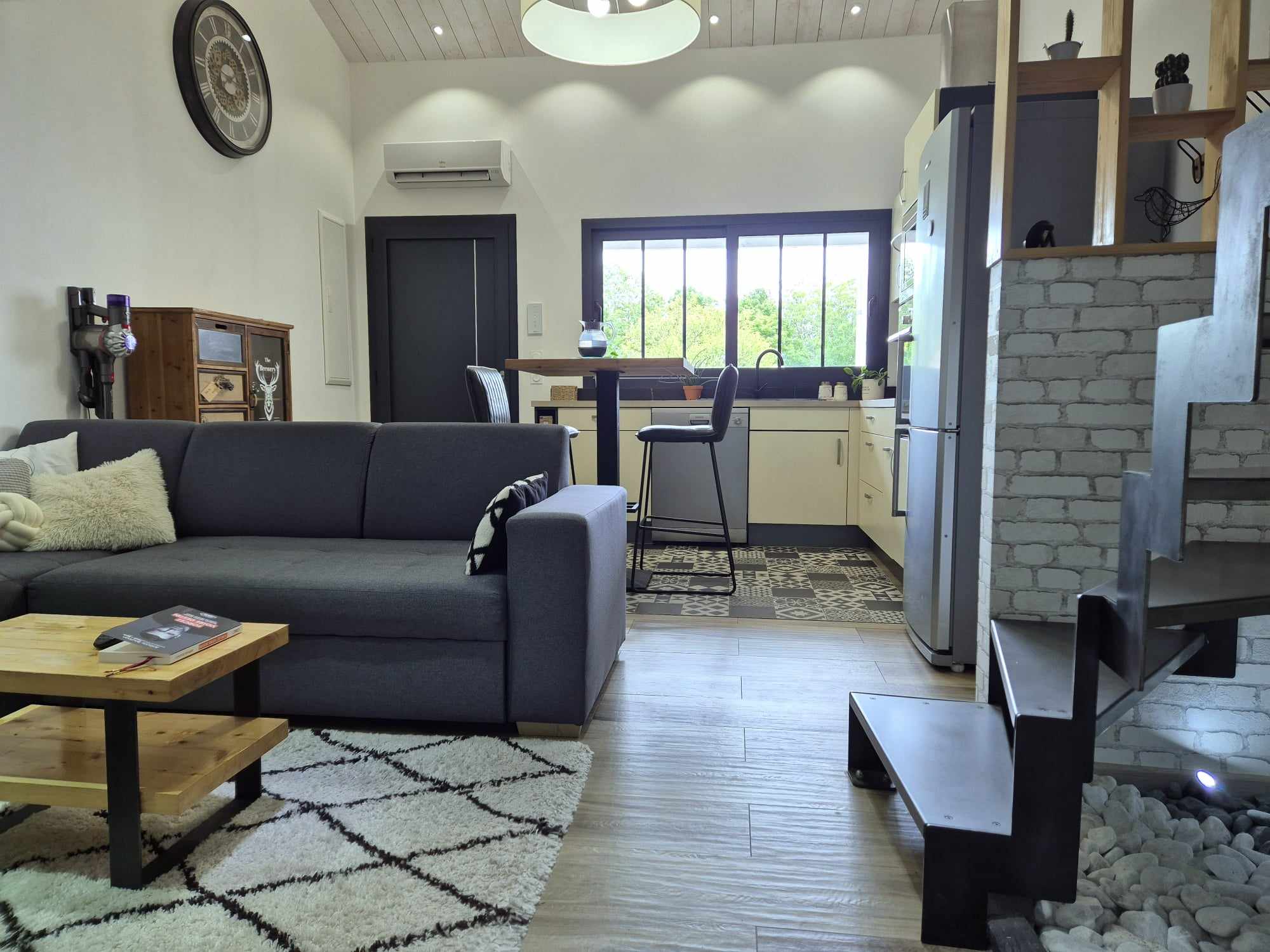 Vue d'un intérieur contemporain à aire ouverte comprenant un canapé d'angle gris anthracite agrémenté de coussins blancs, une table basse en bois clair sur un tapis berbère, et une cuisine équipée en arrière-plan. L'espace se distingue par un escalier industriel en métal noir, un sol en carrelage imitation parquet et des touches de décoration industrielle comme une horloge murale à engrenages.