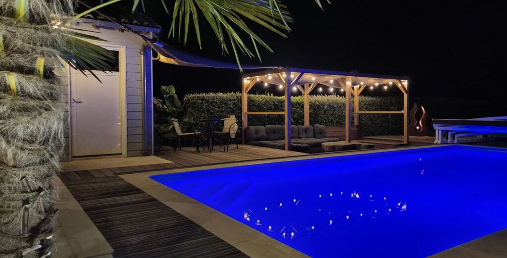Piscine éclairée en bleu la nuit avec une pergola illuminée par des guirlandes ginguettes