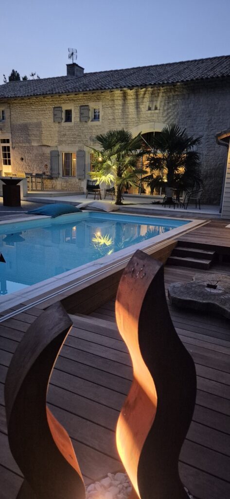 Vue au crépuscule sur une piscine bordée d'une terrasse en bois exotique, une sculpture en corten diffuse une lumière chaude. En arrière plan, une grande maison en pierre est éclairée avec des palmiers