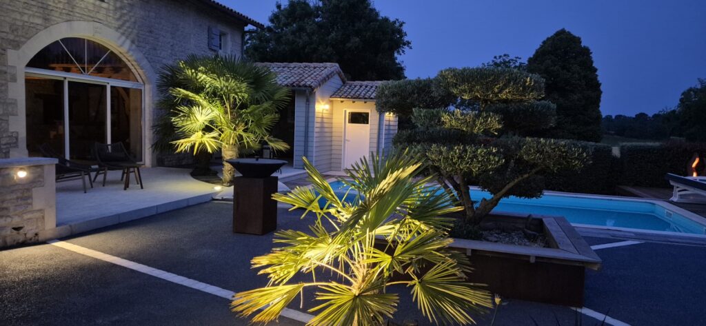 Vue nocturne d'une terrasse élégante avec une piscine éclairée, bordée de palmiers et d'un olivier taillé en nuage. L'espace comprend une maison en pierre avec une grande baie vitrée, un brasero moderne et un éclairage chalereux.
