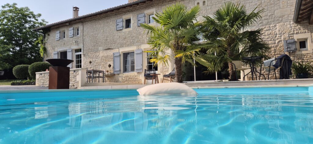 Coussin flottant sur piscine bleu turquoise avec en fond des palmiers, un brasero et une maison en pierre